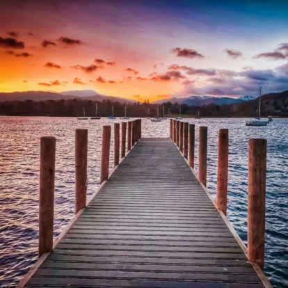 A serene sunset view over a tranquil lake with boats, seen from a wooden pier surrounded by hills.