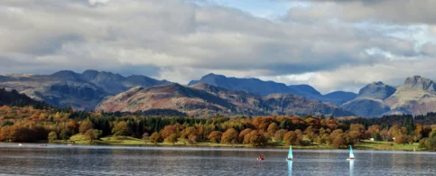 A serene lake with sailboats and lush autumn trees against a backdrop of mountains.
