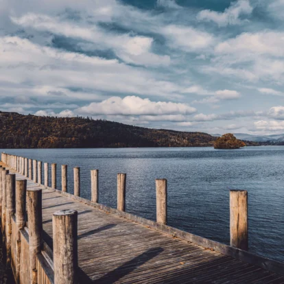 Tranquil lake scene with wooden pier and lush hills in the distance under a cloudy sky.
