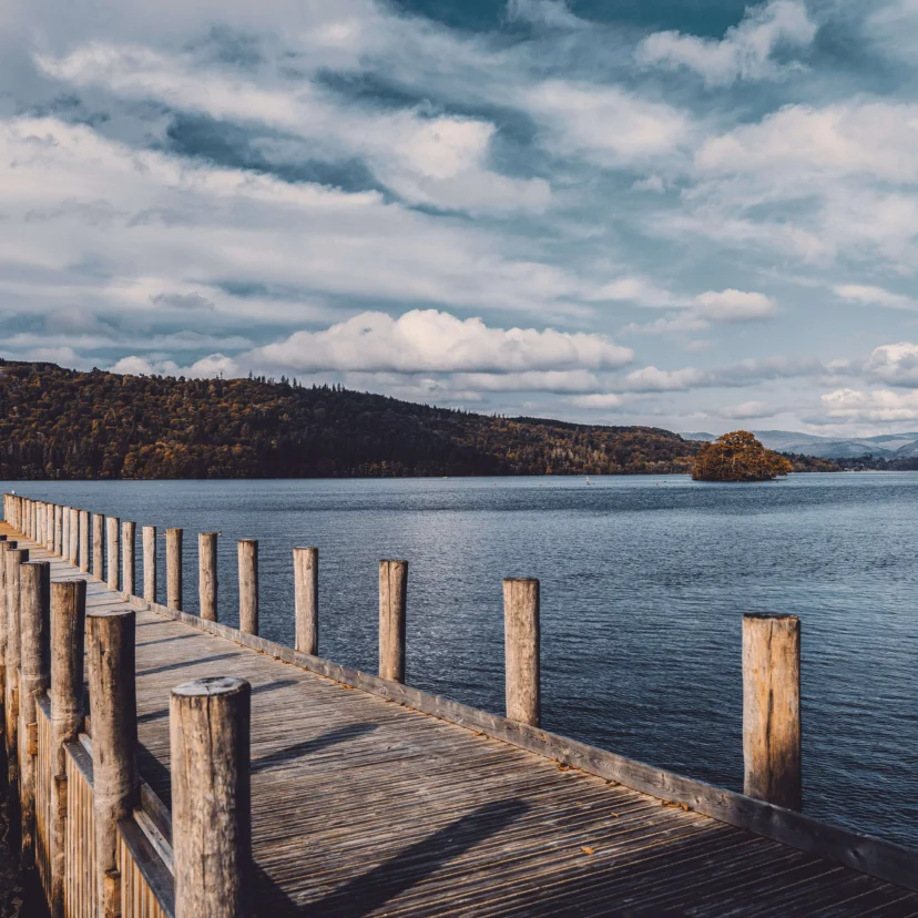 Family walking on a wooden jetty into a calm lake with forested hills and cloudy sky, transcribe: none visible