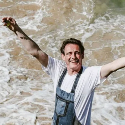 Man joyfully standing on a rocky shoreline with foamy waves and sea in the background.