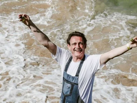 Man joyfully standing on a rocky shoreline with foamy waves and sea in the background.