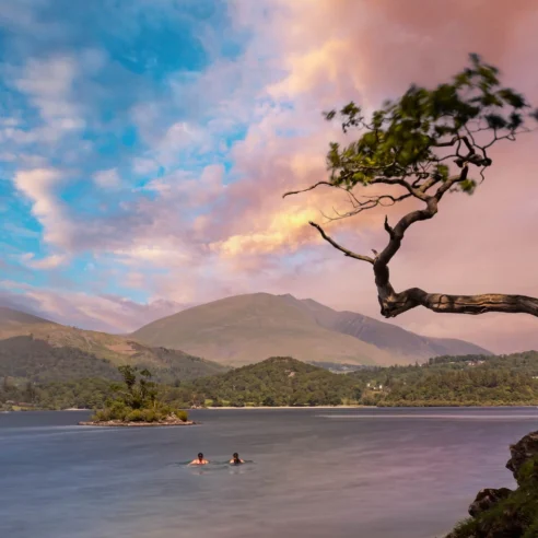 A couple swims in a serene lake, surrounded by majestic mountains and a vibrant sunset sky.