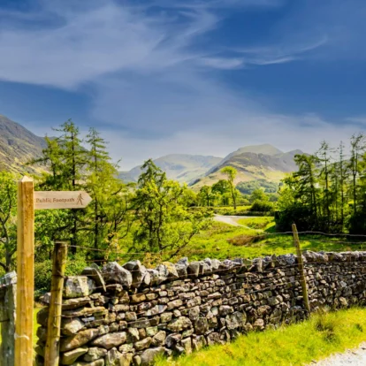 A scenic countryside trail beside a stone wall, leading to green hills under a blue sky.