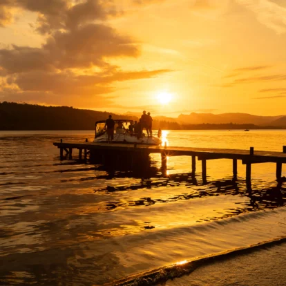 Family walking on pier at sunset, boat docked nearby, tranquil lake setting.
