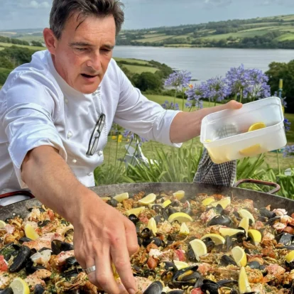 Chef preparing seafood paella outdoors with scenic countryside view in the background.