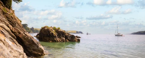 Scenic coastal view with rocky shoreline and sailboats on a calm sea under a cloudy sky.