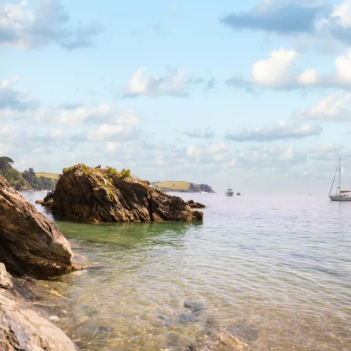Scenic coastal view with rocky shoreline and sailboats on a calm sea under a cloudy sky.