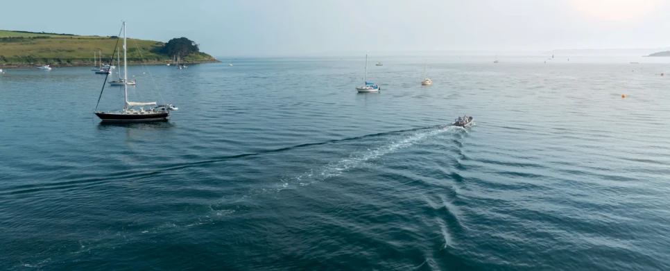 Family on a small motorboat cruising past moored sailboats in a calm coastal bay at sunrise, gentle waves and green headland visible