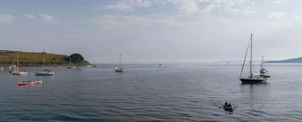 Family rowing and sailing near a calm coastal bay at dawn, anchored yachts and green headland visible