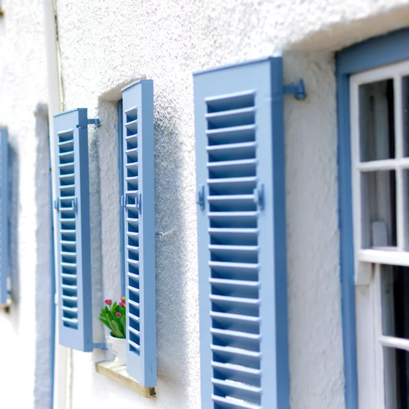 Sunny hotel façade with open blue shutters, white walls and a small flowerpot on the sill