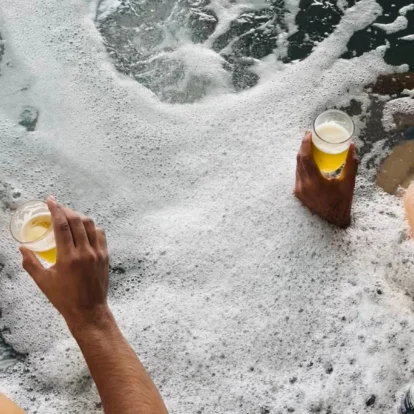 Two people relaxing in bubbly hot tub holding beer glasses, coastal spa setting, casual family moment
