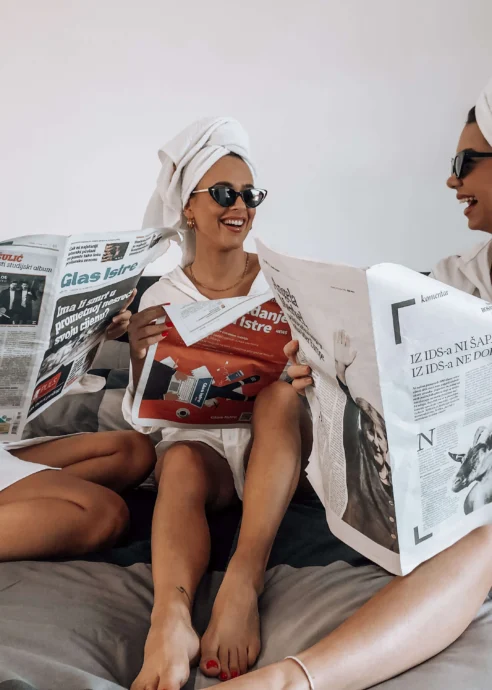 A joyful group of women in towels and sunglasses reading newspapers on a hotel bed.