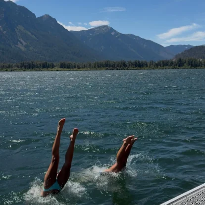 Family members diving from a lakeside dock into clear mountain lake with forested peaks beyond (transcribe: none)